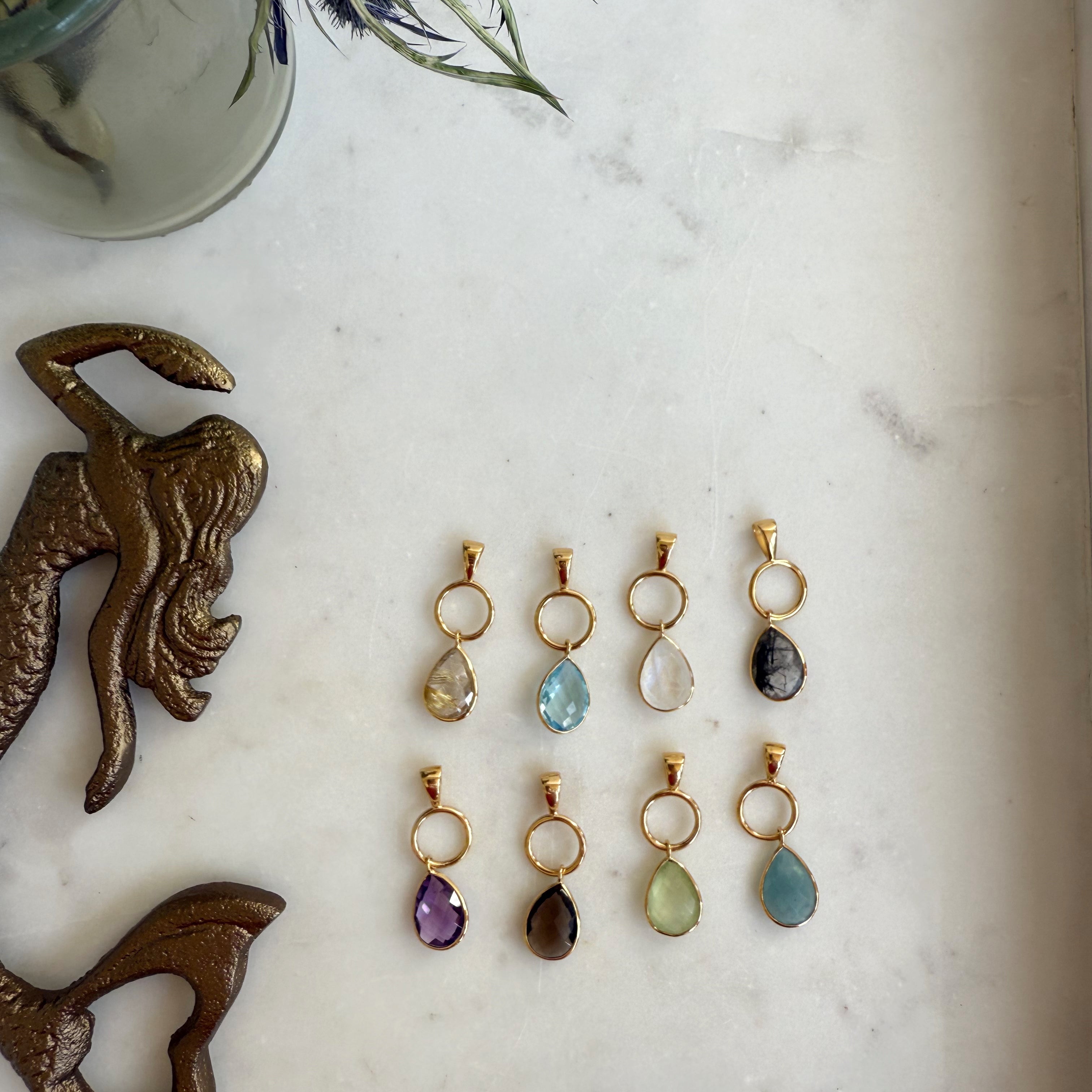 The Mira Drop Pendant by Wild Alabaster — gold-framed faceted gemstone pendants in aquamarine, blue topaz, and golden rutile quartz, photographed on white ceramic dish.